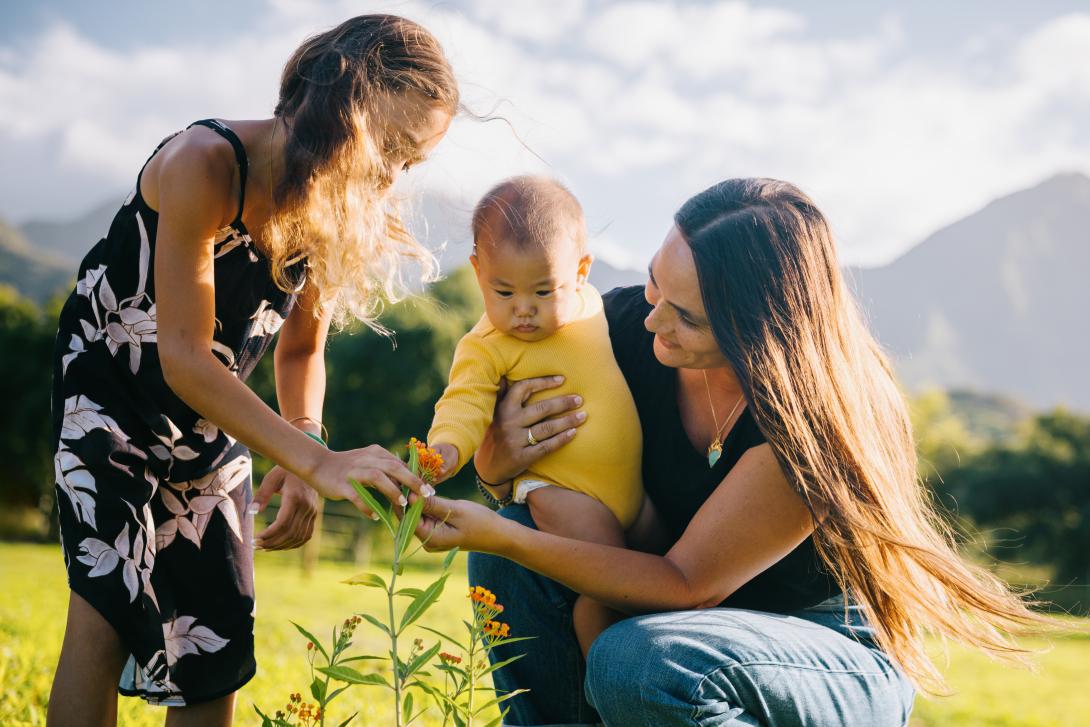 Crouching woman holding infant admiring flower with young girl